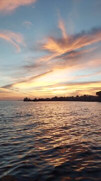 Dynamic vertical short clip of vibrant sunset skies with pink and orange cirrus clouds reflecting across golden ocean ripples near a silhouetted coastal town in Biak, Papua, Indonesia.