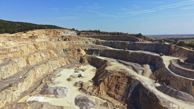 Heavy machinery works at a quarry site, removing minerals from the ground in a hilly area. The sun shines bright as machines dig deep into the earth.