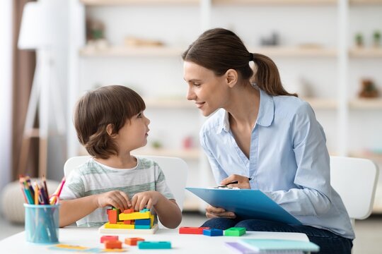Focused Caucasian Mother Engaging In Educational Activity With Young Boy At Home, Smiling While Interacting With Child Creating Colorful Blocks On Table. Family Learning Concept.