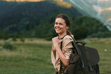 Naklejka premium Young woman traveler with backpack smiles happily standing in green meadow with mountains in background enjoying outdoor lifestyle and nature adventure.