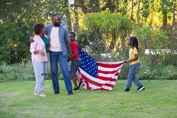 Obraz premium African American family of four walking on grassy lawn in parklike backyard carrying American flag
