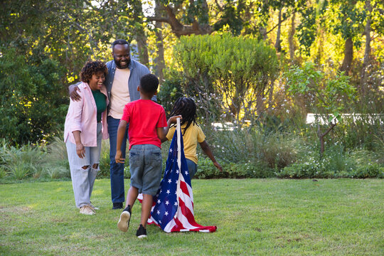 African American family laughing and leaning on backyard lawn while child dragging American flag