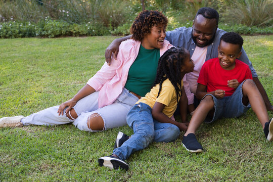 African American family sitting on grassy lawn near shrubs, laughing, wearing clothes and sneakers