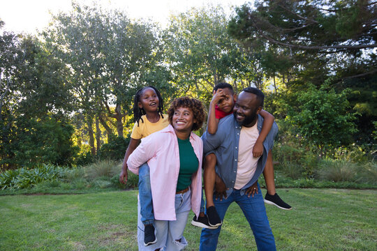 African American family carrying children piggyback and laughing on grassy lawn near trees, shrubs