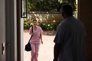 Naklejka premium Asian nurse in pink scrubs walking brick drive to home door carrying black duffel greeting senior