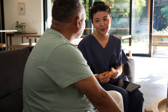 Asian nurse in navy scrubs holding pill-bottle and tablet speaking to client on sofa, copy space