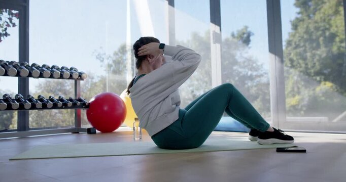 Adult female wearing hoodie doing crunches on yoga mat in home gym, pausing, engaging core training