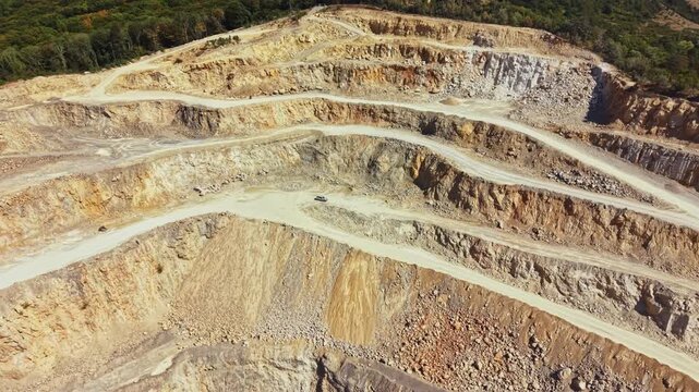 Big machines move dirt and rocks on multiple levels of a quarry. Trucks drive along winding paths to transport materials. Trees surround the area, showing the contrast between nature and industry.