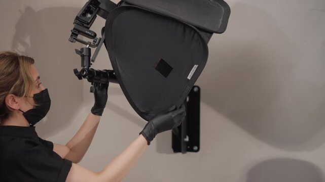 Close up of woman wearing black gloves and mask carefully adjusting professional photography lighting equipment including softbox on tripod against plain wall. Woman is focused as woman