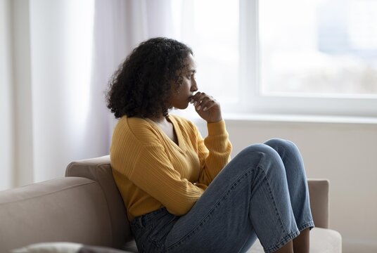 Worried Expression. Black Female Adult, Young Age, Sitting On Couch In Living Room At Home, Staring Out Of Window, Deep In Thought, Reflecting. Concept Of Mental Wellness And Emotional Balance.