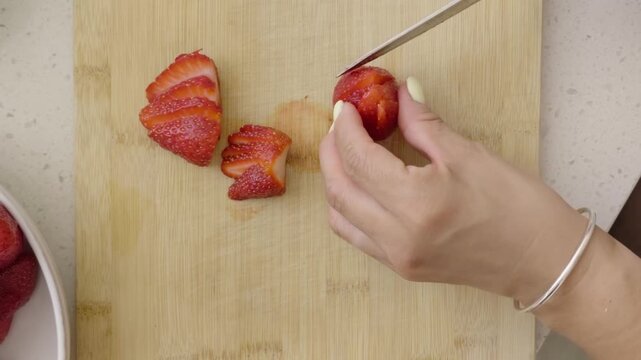 A hand is slicing a fresh strawberry on a light wooden cutting board. The knife is mid‑cut, creating thin, even slices that fan out naturally across the surface. Several already‑cut pieces rest nearby