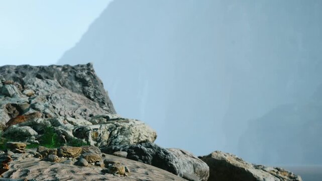 A rocky shoreline is seen at a beach with mist rising from the water. Mountains rise in the background, and the sun is shining on the sand. The scene captures nature in an outdoor setting.