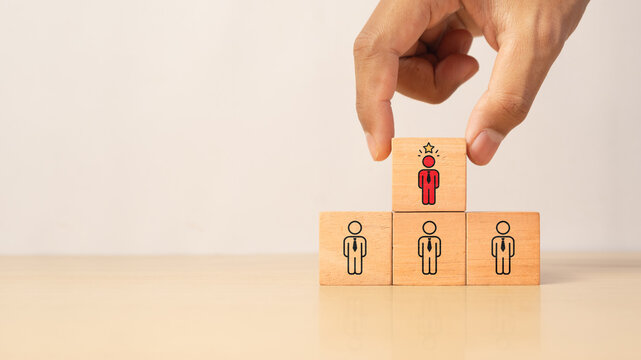 Hand placing a red business person icon on top of a wooden block pyramid, symbolizing leadership promotion, talent selection, employee development, career growth, human resource management.