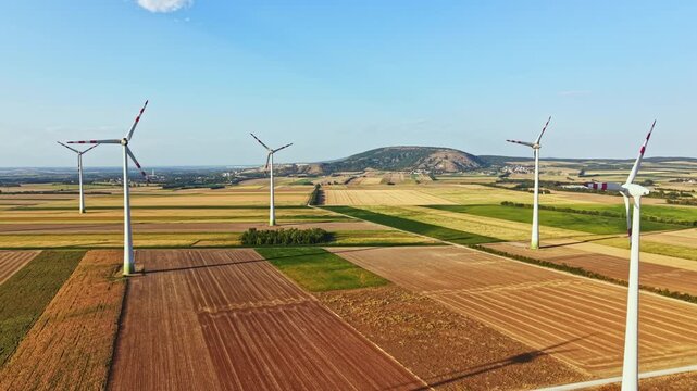 Wind turbines spin in a wide field surrounded by rows of crops and green patches under a clear sky. Hills can be seen in the background near midday.