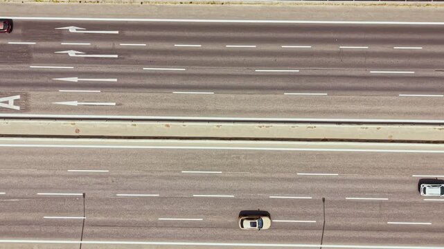 Several cars travel along a busy highway on a sunny day. The view shows lanes marked for traffic with clear white lines. The scene captures vehicles moving in different directions.