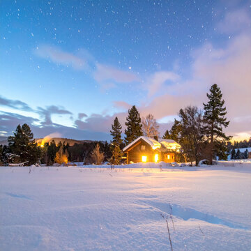 Winter cabin under starry sky with glowing windows, snowy field and evergreen forest create peaceful Christmas scene at blue hour beside mountain landscape