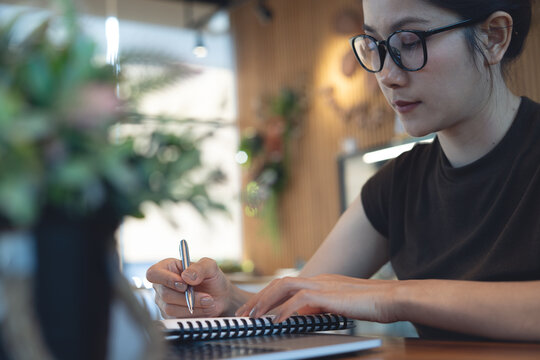 Focused young asian woman with glasses writing in notebook with laptop at desk, remote work and modern professional lifestyle concept.