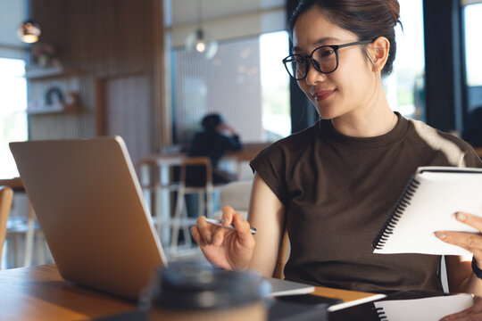 Young asian woman working on laptop and holding notebook planner at cafe in warm natural light, online business and modern remote work lifestyle.