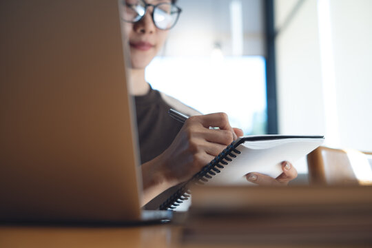 Young asian woman taking notes in notebook while using laptop at cafe in warm natural light, focused productivity and modern remote work lifestyle.