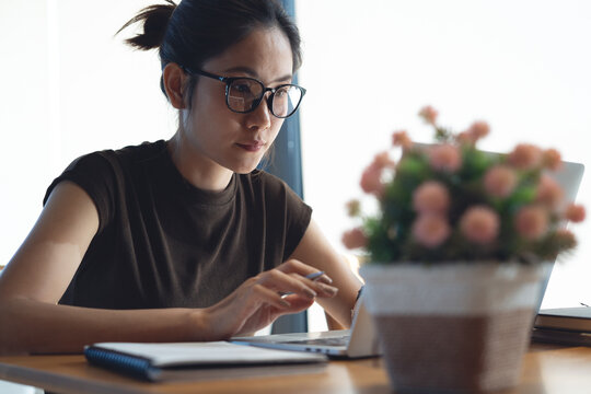 Focused asian woman typing on laptop with notebook on wooden desk in modern workspace in natural light, remote work, online business concept.