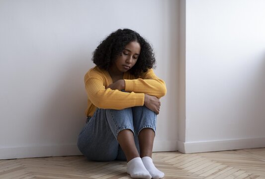 Worried Young Black Woman Sitting On Floor With Arms Wrapped Around Knees, Appearing Deep In Thought, In A Bright Indoor Setting. Emphasizing Mental Health Awareness And Emotional Wellbeing.