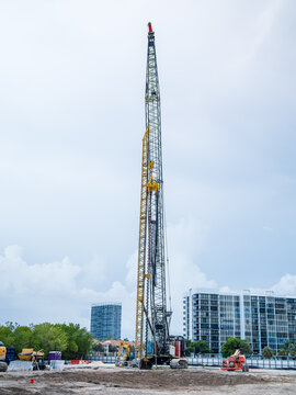 Tall heavy-duty drilling rig machine operating on a sandy construction site with residential high-rise buildings in the background.