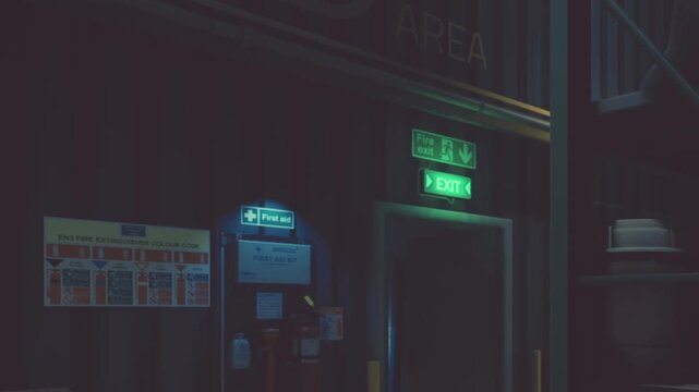 A storage area shows a first aid kit mounted on the wall. A bright exit sign is visible nearby. Shelves line the walls, holding various items. The lighting is dim but clear.