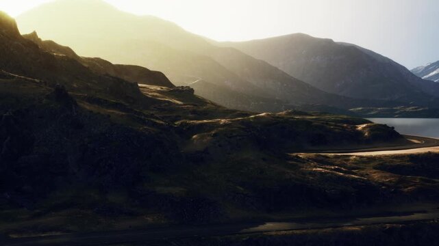 A landscape shows mountains, water, and a winding road at sunrise. The sun creates a warm glow on the hills. The scene captures the natural beauty of the outdoors.