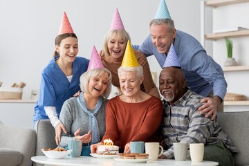 Naklejka premium Joyful Emotion. Group Of Older Women And Men, Senior Caucasian And Black, Celebrating Birthday Together In Cozy Living Room, Smiling Around Cake With Candles. Family Celebration Concept.