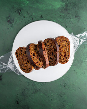 Sliced rye bread loaf in transparent plastic packaging on round white plate over textured green background. Top down flat lay view for bakery food marketing retail commerce and grocery store branding.