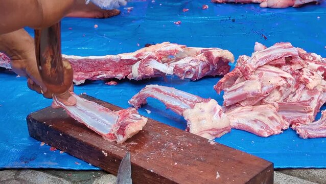 A close-up of a butcher using a large blade to slice fresh raw meat and ribs on a wooden cutting board over a blue tarp.