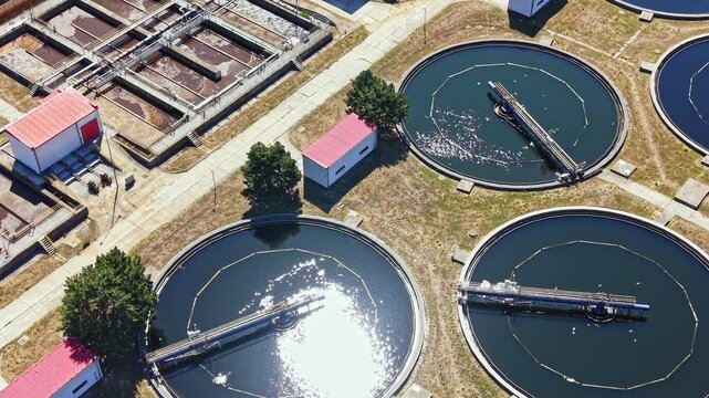 Water flows through round tanks at a wastewater treatment facility. Buildings surround the tanks under a clear sky. The scene shows a vast open area with a structured layout.