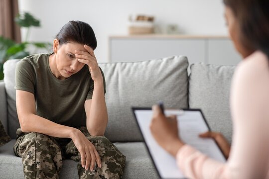 Worried Latina Woman Sitting On Couch Speaking With Female Therapist In Office, Seeking Help And Support For Mental Health Issues, Engaging In Counseling Session In Professional Setting.