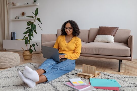 Focused Black Woman Sitting On Floor In Cozy Living Room, Working On Laptop With Books And Stationery Nearby, Engaged In Productive Activity At Home. Work From Home Concept.