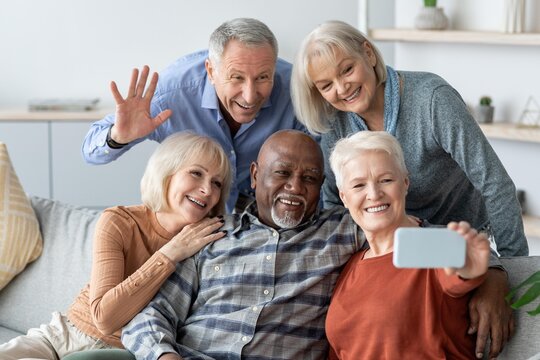 Happy Multiracial Group Of Five Seniors, Including Two Males And Three Females, Smiling And Posing For Selfie Together At Home, Celebrating Friendship And Joyful Moments In Cozy Living Room.