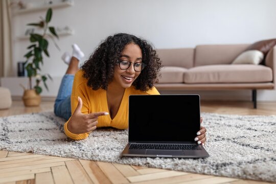 Smiling Young Black Woman Pointing At Empty Laptop Screen While Lying On Carpet At Home, Engaged In Online Activity, Enjoying Technology And Digital Life. Concept Of Remote Work And Lifestyle.