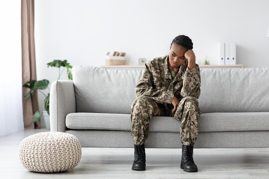 Worried Young Black Female Soldier Sitting Alone On Couch At Home, Resting Her Head On Hand, Contemplating Her Thoughts. Indoor Setting Reflecting Mental Health Concerns And Emotional Struggles.