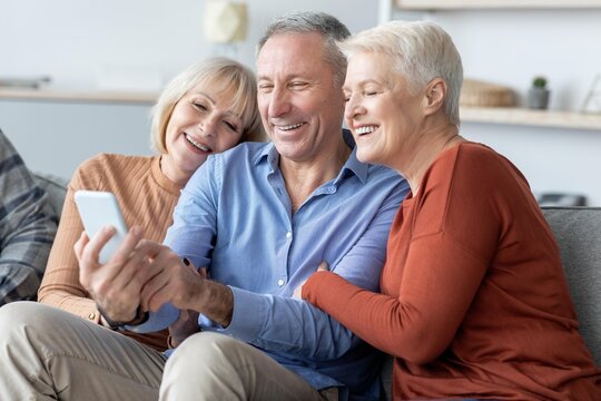 Smiling Caucasian Seniors Enjoying Quality Time Together, Engaged With Smartphone While Sitting Comfortably In Their Living Room At Home. Family Bonding And Connection Concept.