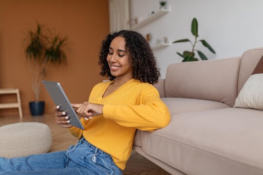 Smiling Black Woman In Her 20s Using Tablet While Relaxing On Couch At Home, Engaged And Focused On Screen. Cozy Indoor Setting Portraying Modern Lifestyle And Comfort.