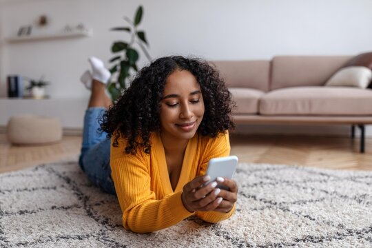 Smiling Young Black Woman Relaxing While Lying On Carpet At Home, Using Smartphone To Browse Social Media Or Chat, Enjoying Free Time And Comfort. Indoor Lifestyle Concept.