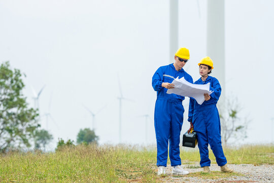 Two renewable energy engineers wear coverall inspecting blueprint at wind turbine field. Male and female hold toolbox inspection engineer team working at windmill site for sustainability project.