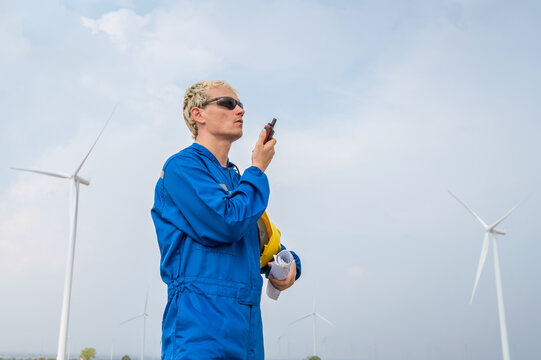 Male engineer in coverall looking and talking by radio communication working at wind turbine farm renewable energy Technician man holding yellow hardhat blueprint inspecting wind power plant