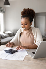 Fototapeta na wymiar Smiling young woman uses calculator seated at table surrounded by bills, receipts, and laptop, busy in budgeting or personal finances management in cozy living room. Financial awareness, money control