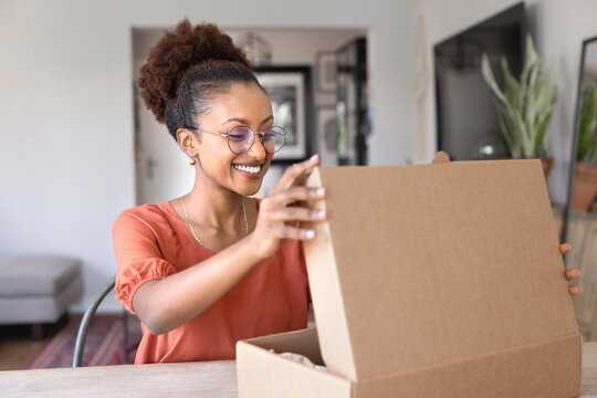 African woman sit at table in living room opens cardboard parcel box, smiling with excitement and curiosity, enjoy received gift from friend or family. Online shopping satisfaction, delivery services
