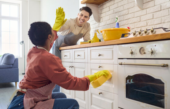 Smiling couple cleaning kitchen with gloves and sponge. They wipe cabinets and oven fronts, share a high five, keeping chores efficient in aprons at home. Teamwork and joyful home care.