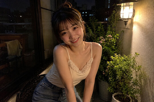壁灯のある夜のベランダで前かがみに笑うクリーム色キャミソールの女性