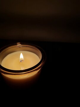 Close-up of a lit scented candle with a glowing flame inside a clear glass jar set against a dark, moody background.
