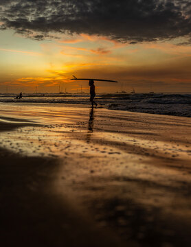 Surfer silhouette walking on a tropical beach carrying a surfboard at dramatic sunset