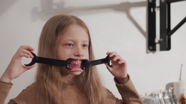 Close up of young girl holding black dental retractor to show orthodontic braces in mouth. Girl with long hair wears brown top background includes medical equipment and white wall. Soft