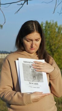 Distraught and worried young woman standing outdoors in a park, holding a stack of reward flyers with a photo of her beloved missing dog, her face filled with despair and sorrow